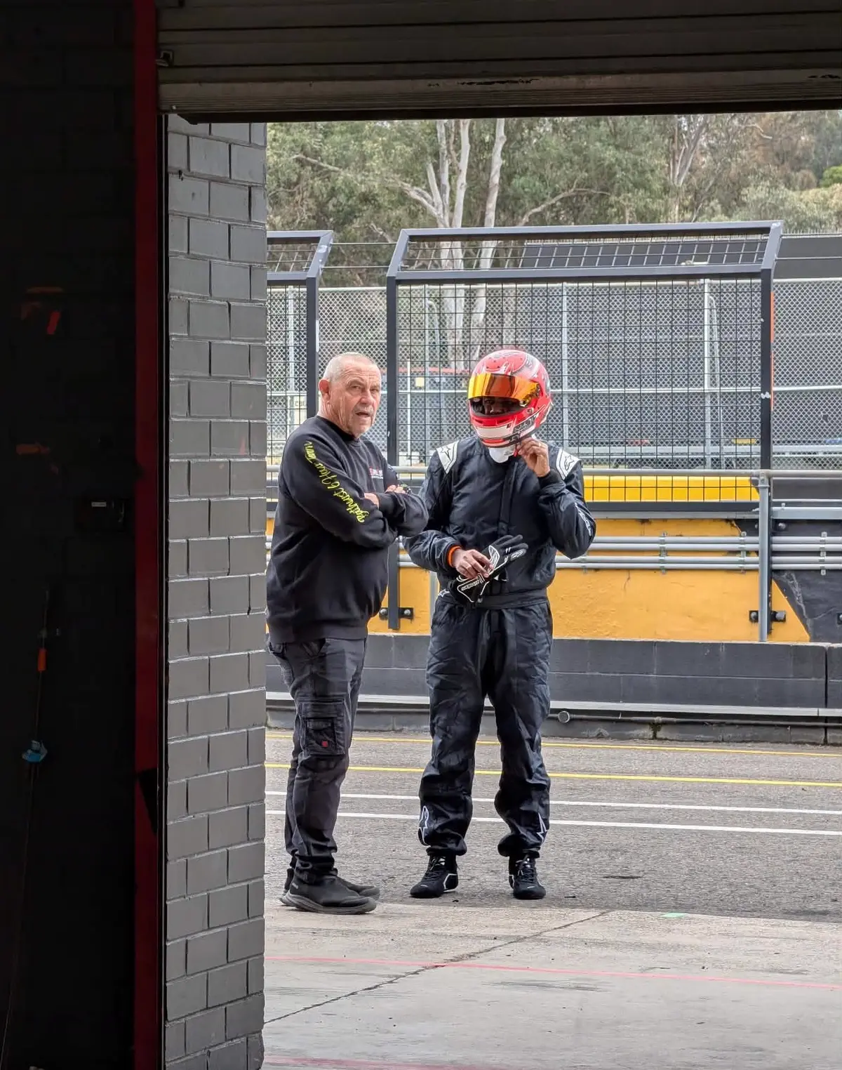 Nikunj in a race suit and helmet in the pit lane, talking with another person in team clothing.