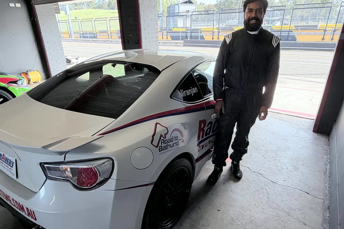 Nikunj in a race suit beside a Toyota 86 in a pit garage, his surname on the rear quarter window and the circuit visible through the open roller door.